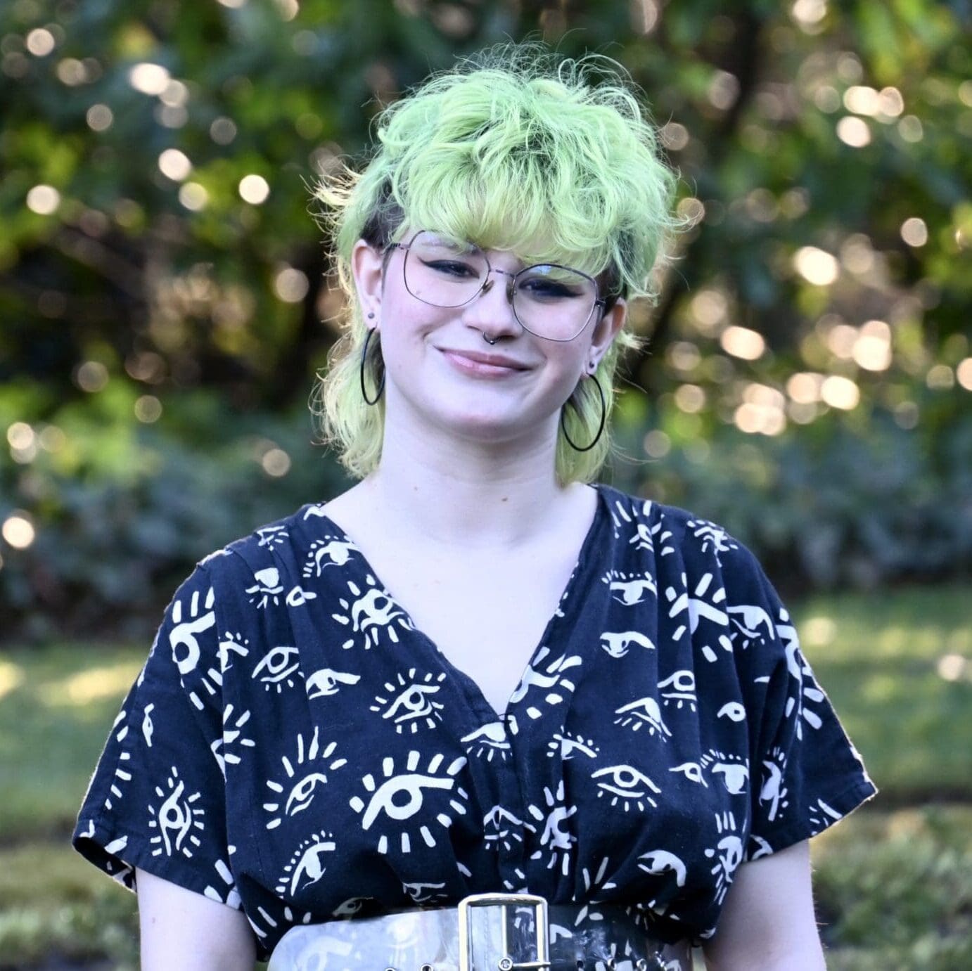 Venus Edlin, a white androgynous person, smiles in a headshot. They have green hair, wear glasses and are donning a jumpsuit with an eye motif.